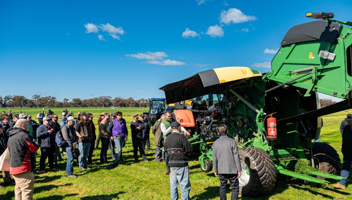 A group of people stand on a grassy field listening to a presenter explain the workings of a large green agricultural machine with panels open to reveal the engine. The sky is bright blue with scattered clouds.