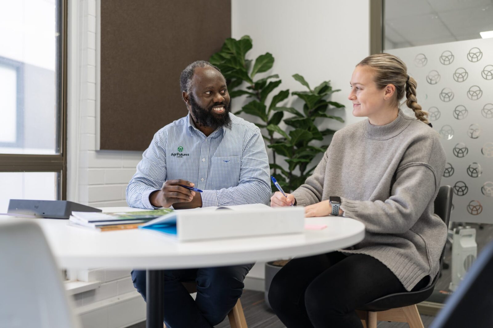 Two colleagues seated at a small round table in a modern office, reviewing documents and taking notes, with folders and papers spread across the table and indoor plants in the background.