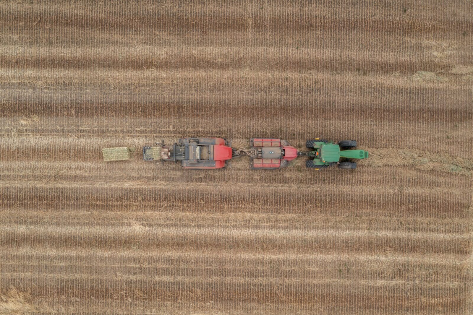 Aerial view of a green tractor pulling a red baler across a harvested field, creating rectangular hay bales in long, parallel rows of stubble.