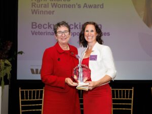 Becky Dickinson stands on stage receiving the Victorian AgriFutures Rural Women’s Award, holding a glass trophy alongside the presenter, with an event screen displaying the Award title behind them.